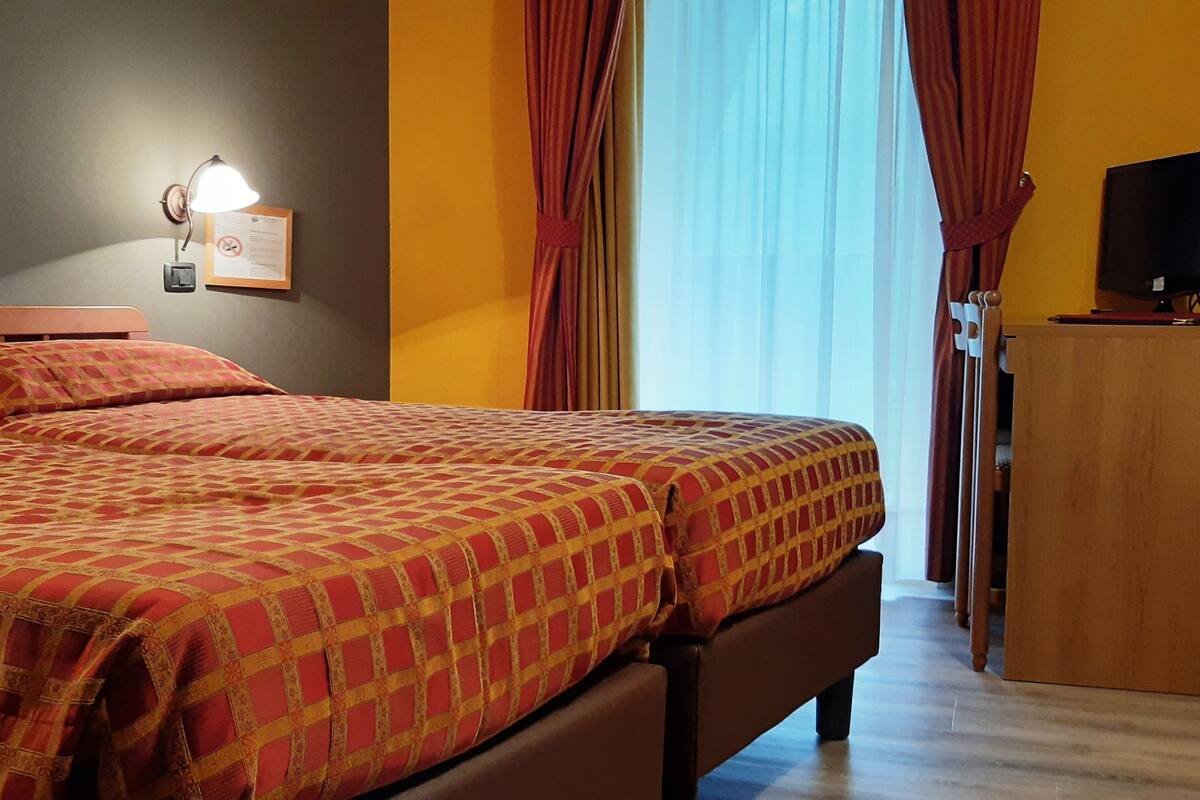 A hotel room with two beds, a desk, a television, and a wall-mounted lamp.