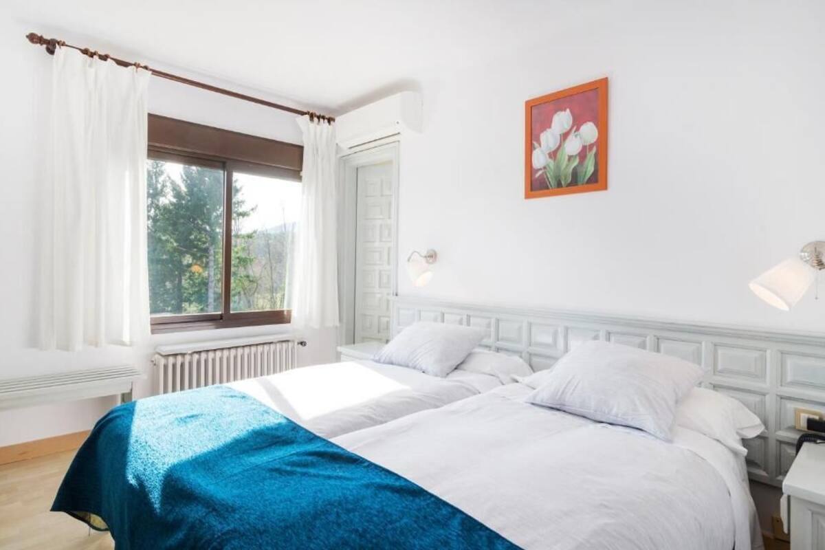 Quarto com duas camas, roupa de cama branca, manta azul, janela com cortinas brancas, radiador, luz acoplada à parede e quadro com flores de tulipa.