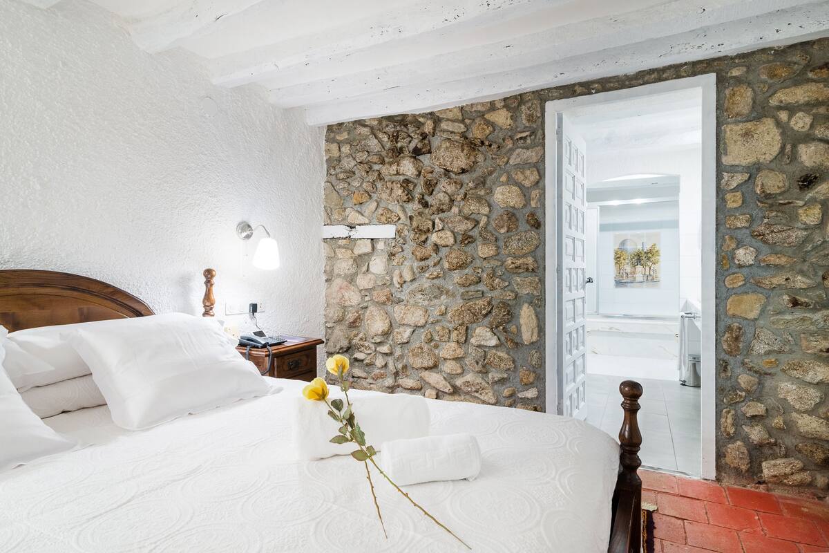 Quarto com parede de pedra, estrutura de cama de madeira, mesa de cabeceira e porta branca que dá para outro cômodo.