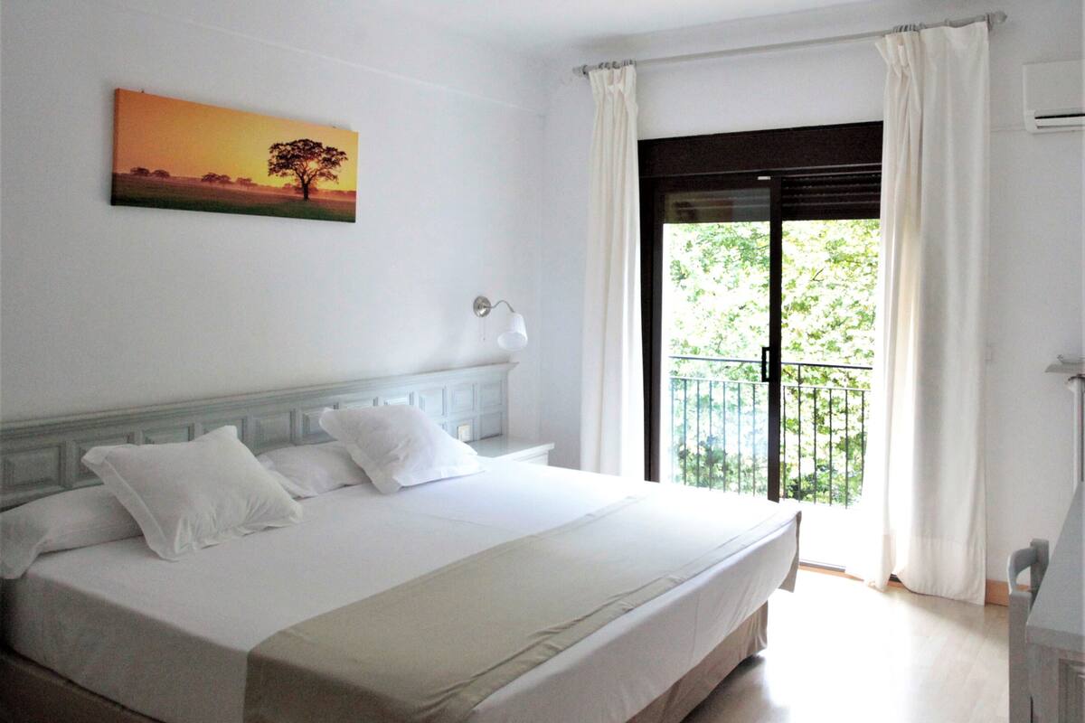 Quarto de hotel com uma cama grande, um quadro na parede, uma janela com cortinas e uma varanda com vista para árvores.