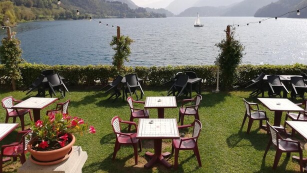 An outdoor seating area with tables and chairs overlooking a lake, surrounded by greenery and mountains.