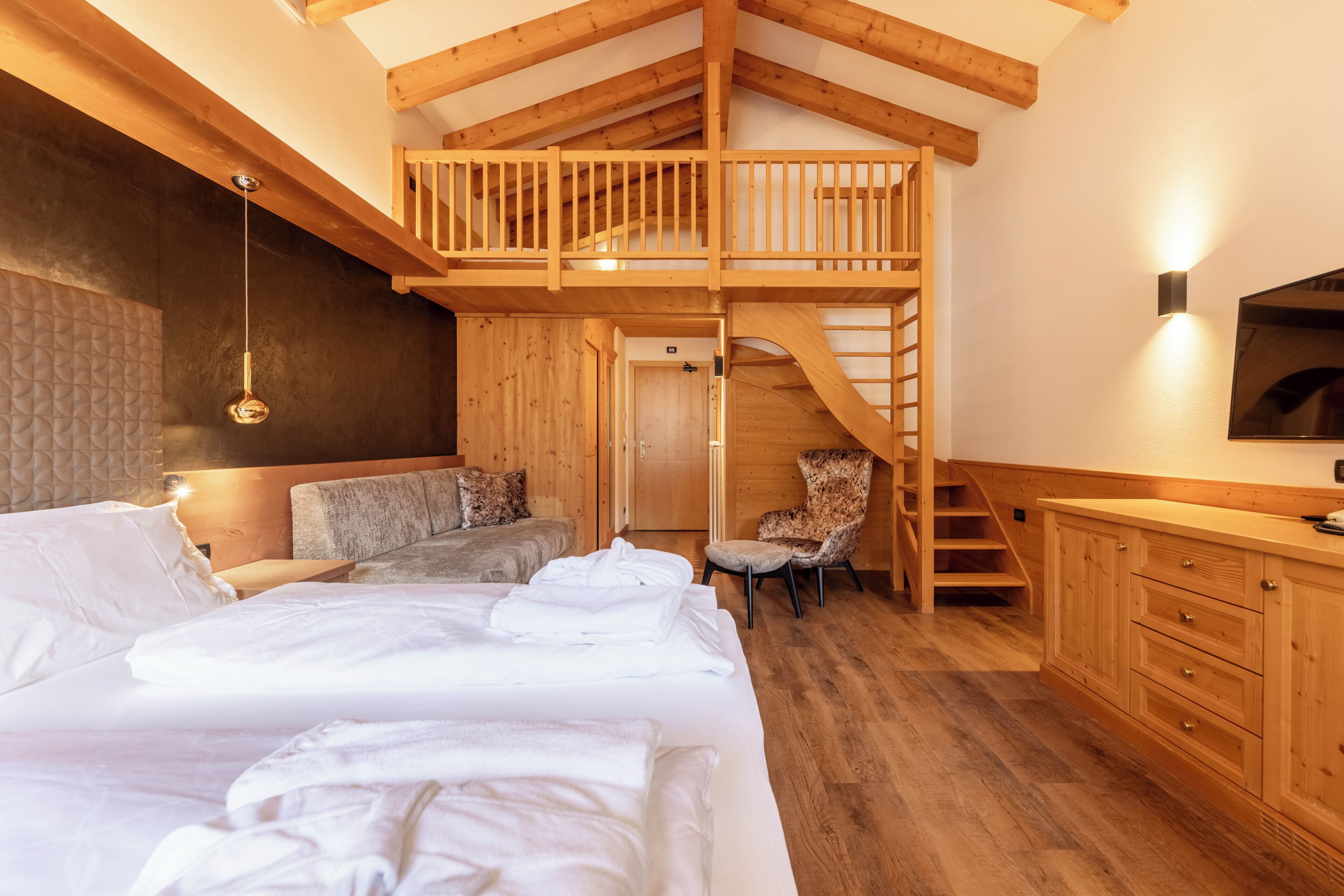 A bedroom with a loft, wooden beams, a bed, a chair, a TV, and a staircase.
