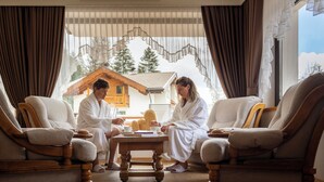 A room with two wooden armchairs, a wooden coffee table, and a view of a wooden cabin through sheer curtains. - Hotel Terme Antico Bagno (San Giovanni di Fassa)