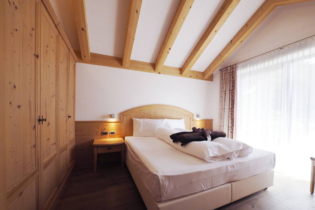 A bedroom with a wooden ceiling, a bed with white bedding, a nightstand with a lamp, and a window with curtains.