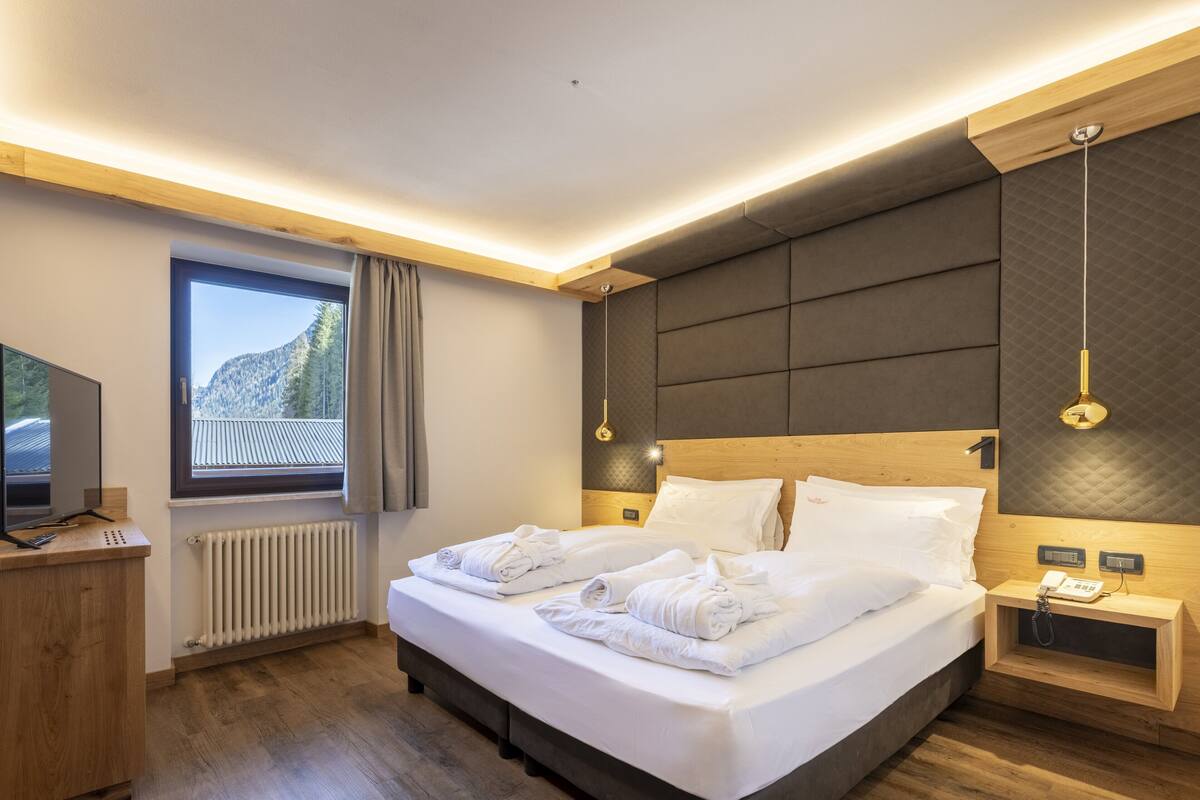 A modern hotel room with a large bed, a desk with a TV, a window with a view of mountains, and a radiator.