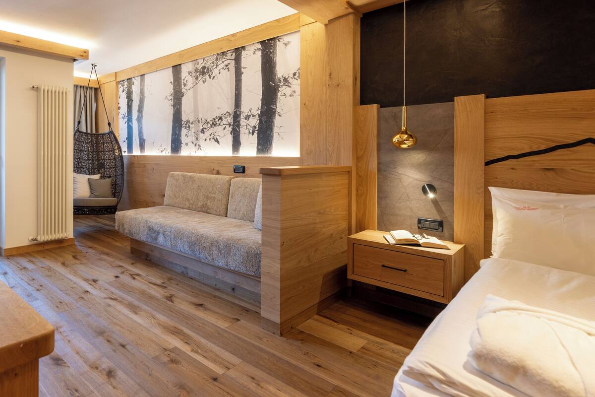 A room with a wooden floor, a bed with white bedding, a hanging chair, a nightstand with a lamp, and a wall with a tree painting.