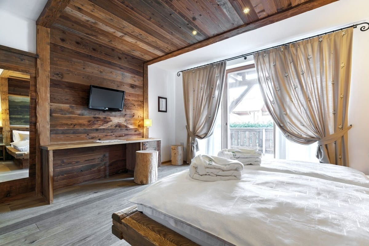 Une chambre avec un plafond en bois, un lit recouvert de draps blancs, un téléviseur fixé au mur et des rideaux à la fenêtre.