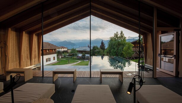 A modern room with a large glass window, wooden ceiling beams, and a view of a swimming pool and mountains. - Pineta Hotels Nature Wellness Resort (Predaia)