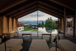 A modern room with a large glass window, wooden ceiling beams, and a view of a swimming pool and mountains.