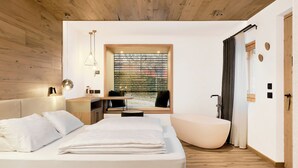 A modern bedroom with a large bed, a sitting area, a bathtub, and a window with a view. - Pineta Hotels Nature Wellness Resort (Predaia)