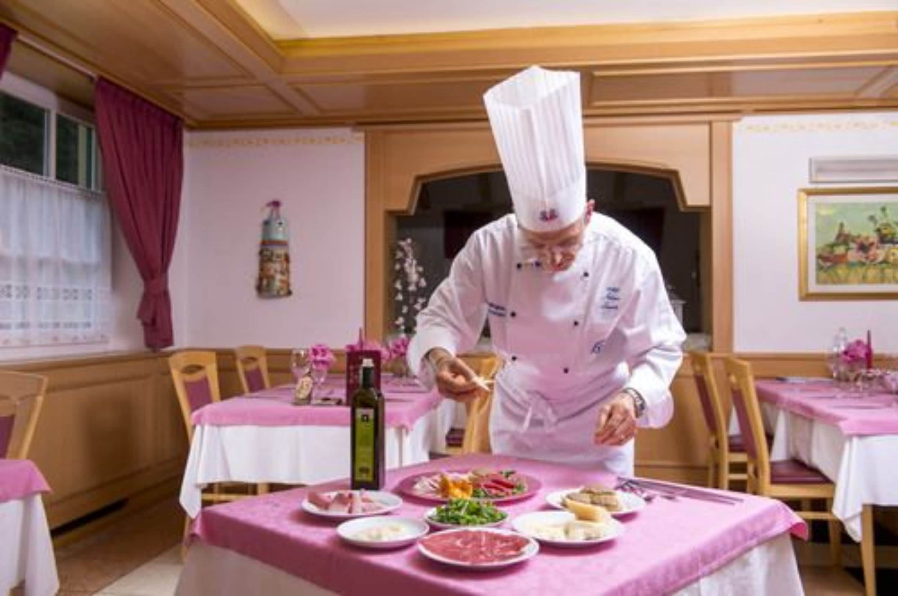 A chef preparing food in a dining area with pink tablecloths and wooden chairs.
