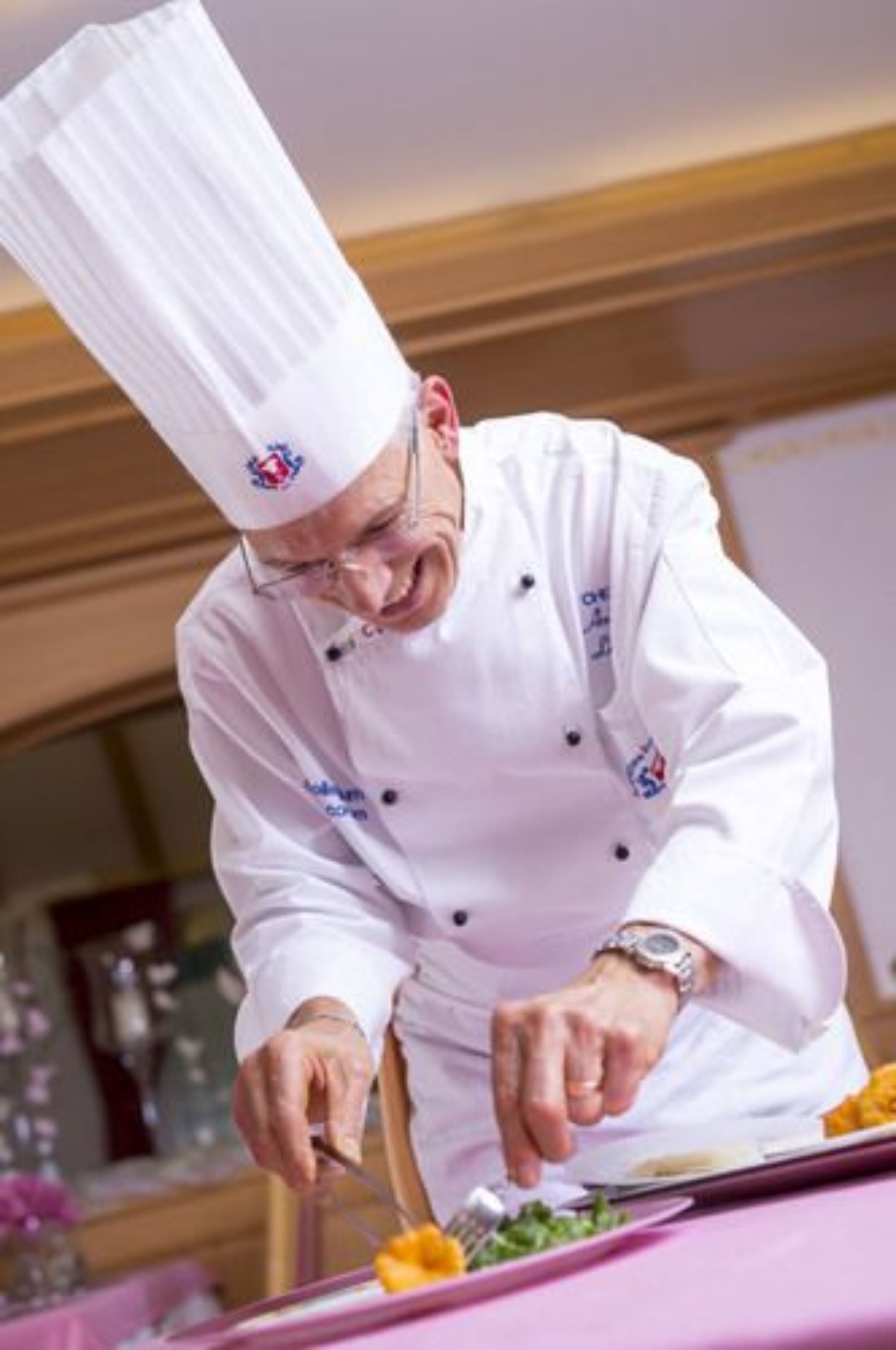 A chef in a white uniform with a tall hat, preparing food on a table with a pink tablecloth.