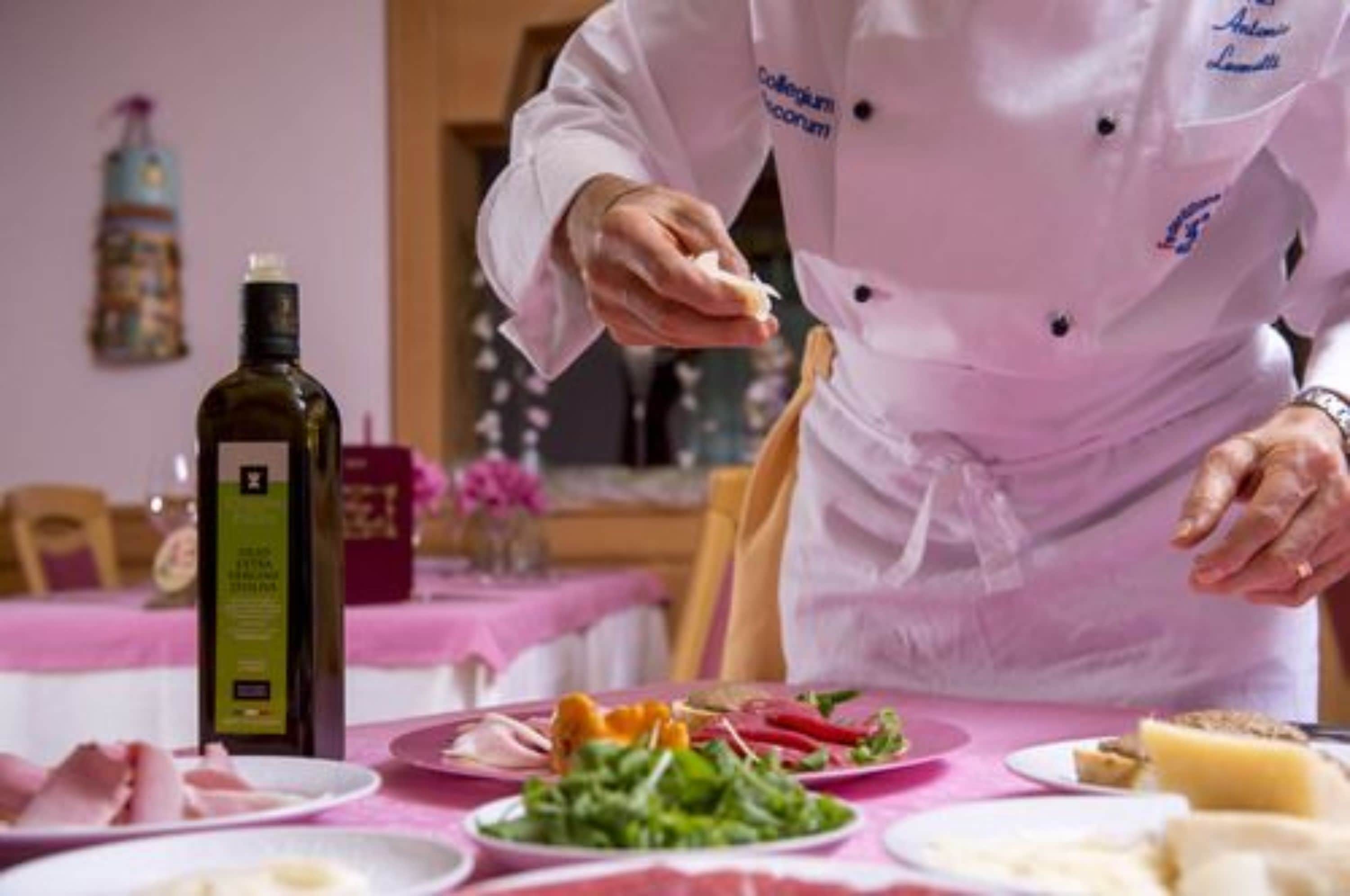 Um cozinheiro com uniforme branco e etiqueta de identificação, preparando comida em uma mesa com diversos pratos, incluindo uma garrafa de azeite, legumes e carne.