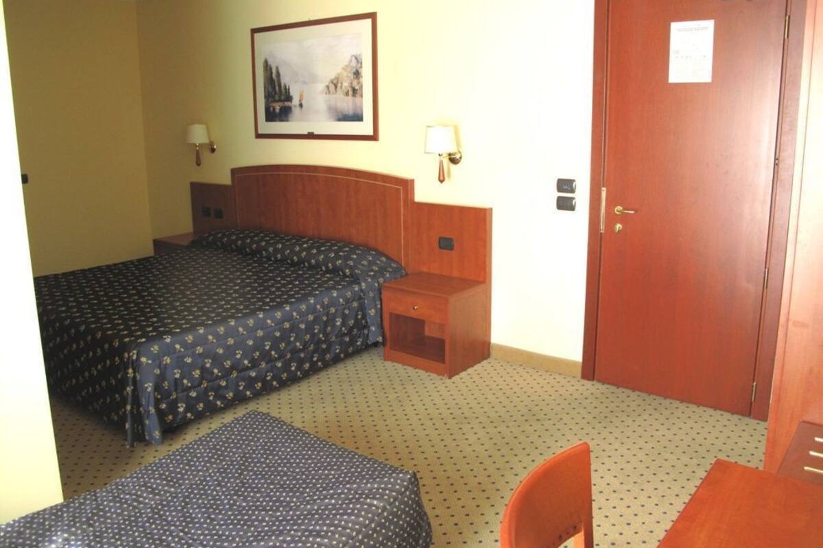 A hotel room with two beds, a wooden headboard, a small bedside table, a chair, a door, and a framed picture on the wall.