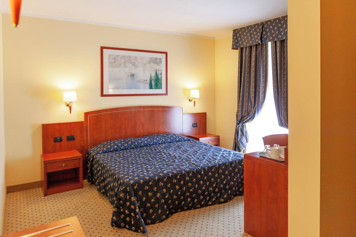 A hotel room with a large bed, two bedside tables, a nightstand, a desk with a chair, a framed picture on the wall, and a window with curtains.