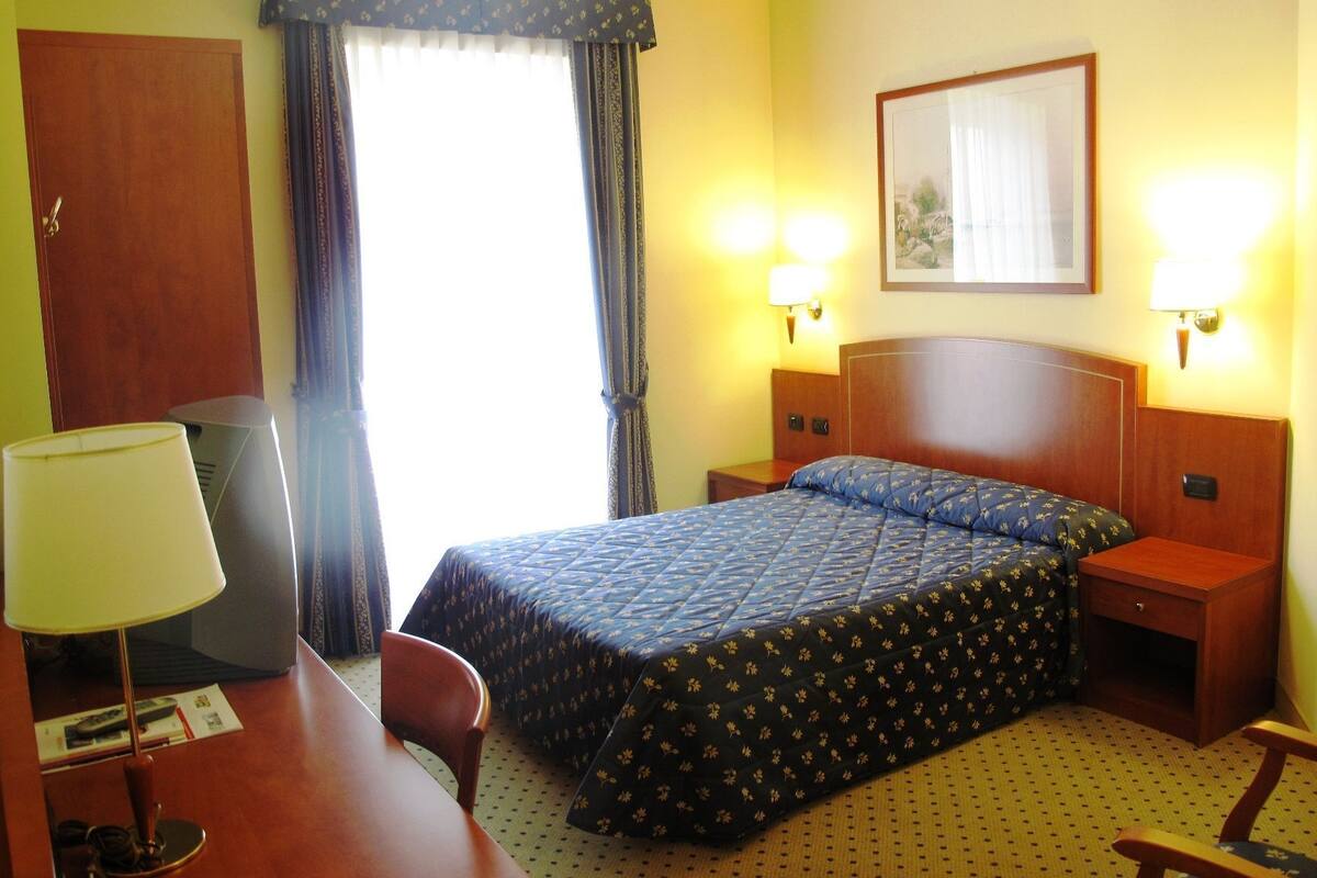 A hotel room with a bed, bedside tables, a desk with a lamp, a chair, and a window with curtains.