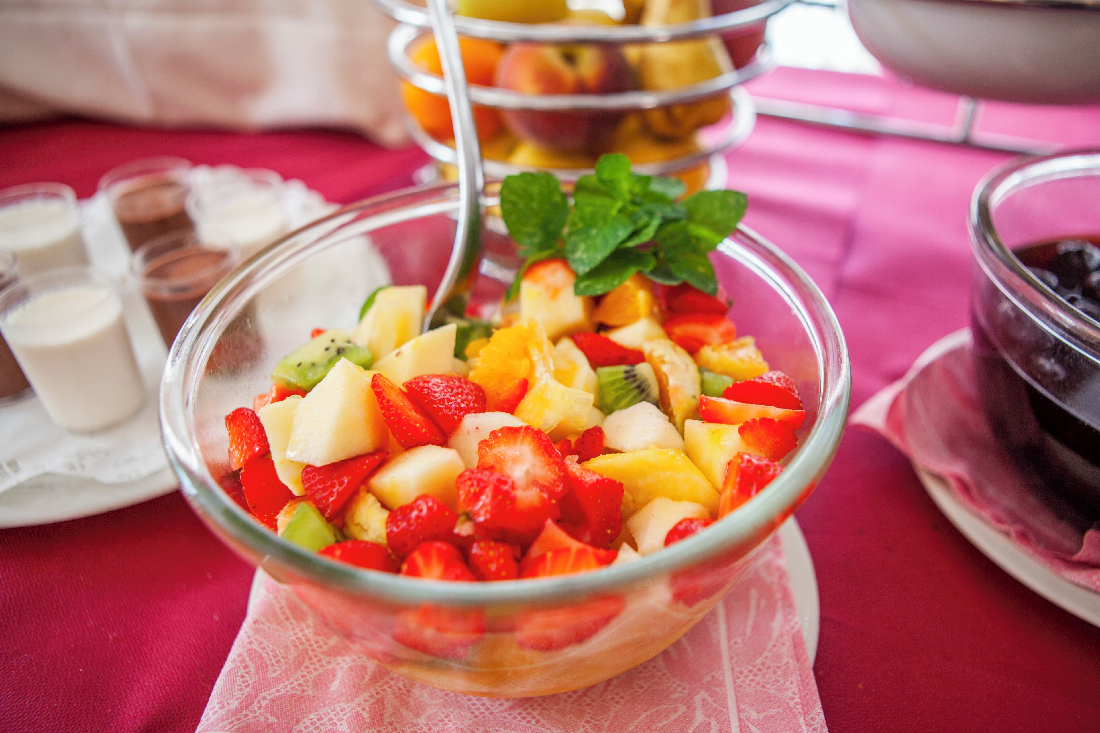 Ein Obstsalat in einer Glaskugel, garniert mit Minze, umgeben von weiteren Schüsseln und Tassen auf einer Tischdecke.