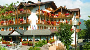A multi-story hotel with balconies adorned with flowers, a visible entrance with a sign, and a mountainous backdrop. - Olisamir (Cavedago)