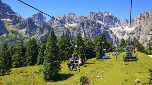 A ski lift with passengers, surrounded by green trees and rocky mountains in the background. - Olisamir (Cavedago)