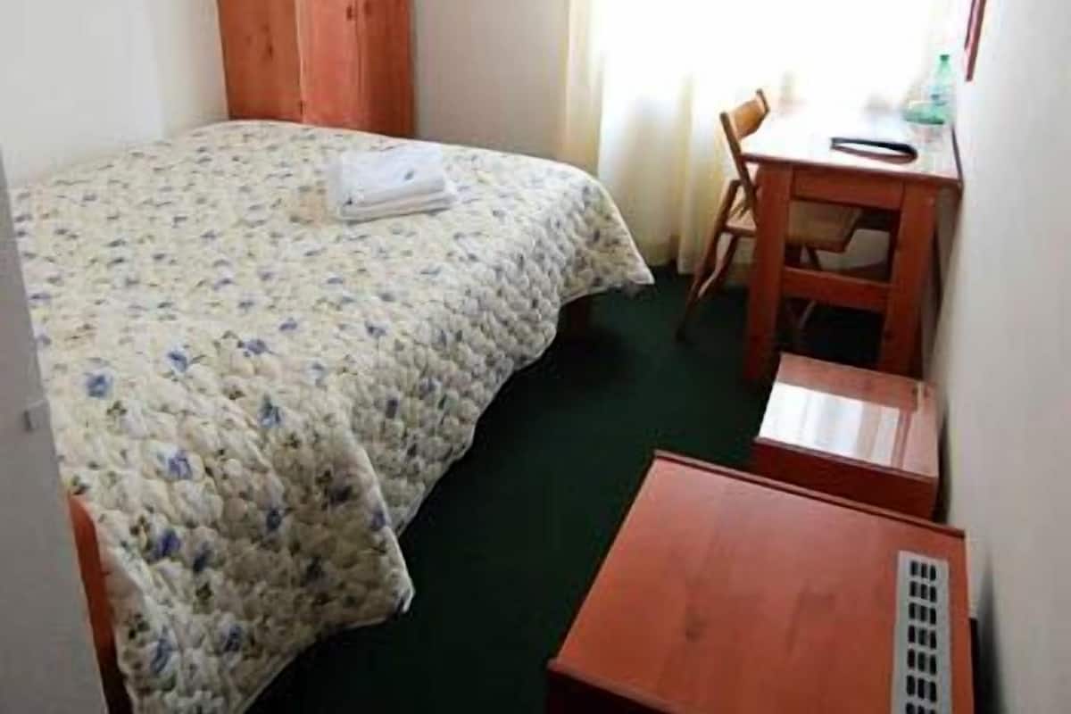 A small, simple room with a bed, a wooden wardrobe, a desk with a chair, and a window with curtains.