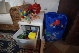 Un soggiorno con un divano bianco, una poltrona in vimini, un contenitore di plastica, una borsa blu e una piccola struttura di legno per giocattoli.
