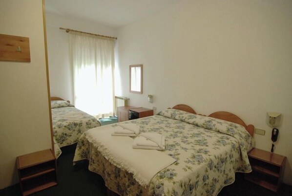 Quarto de hotel com duas camas, uma pequena escrivaninha, uma cadeira, um telefone e uma janela com cortinas.