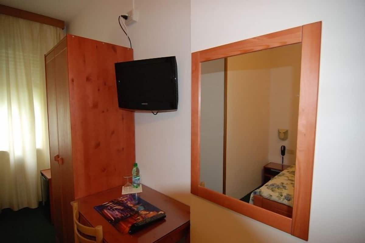A small room with a wooden desk, a television mounted on the wall, a bed with a wooden frame, and a mirror reflecting the room.