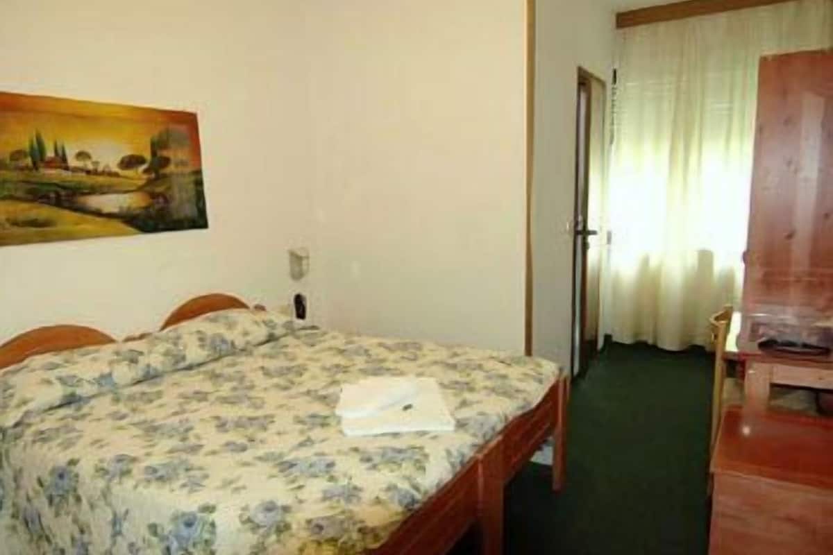 A hotel room with a bed, a painting on the wall, a wooden desk, and a door leading to another room.