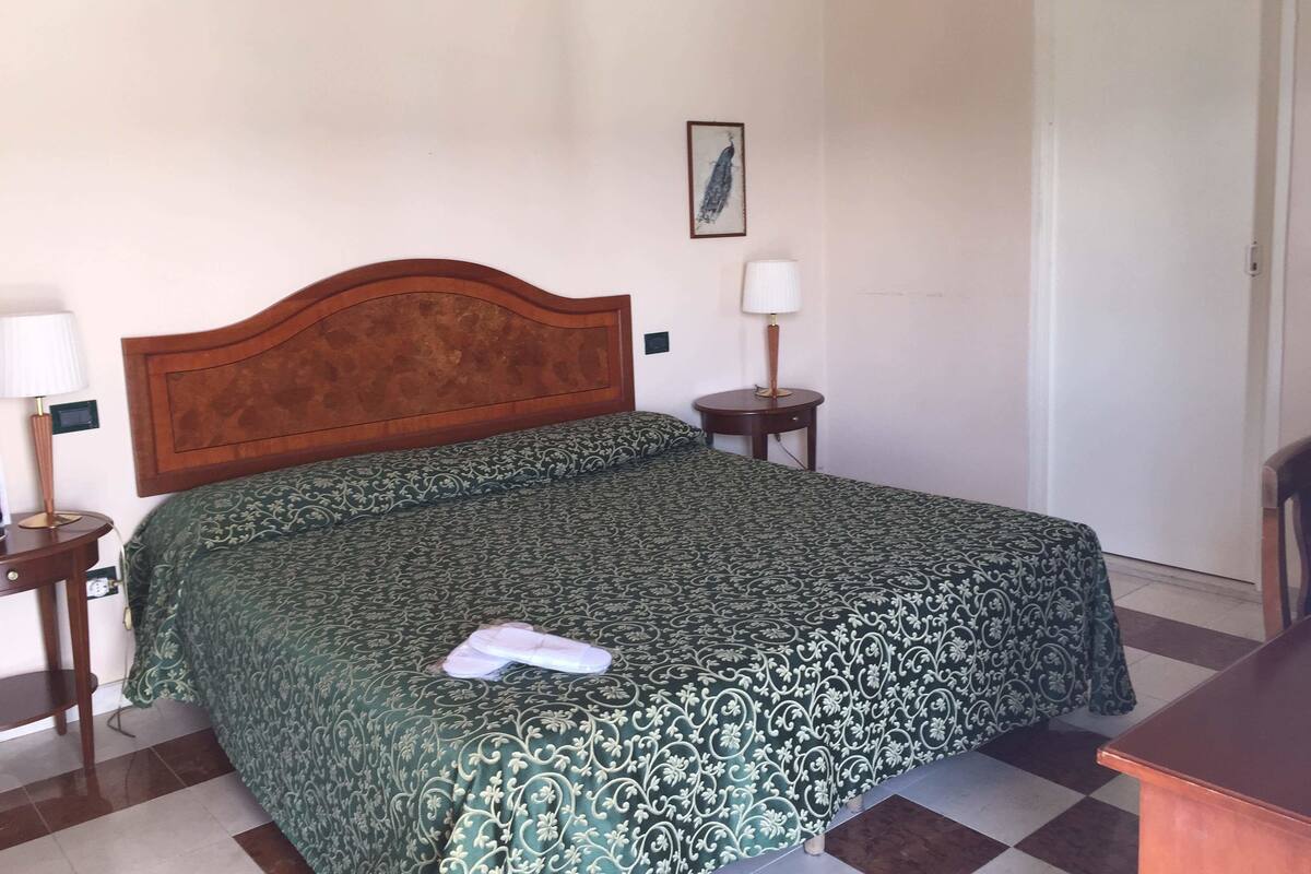 Una habitación de hotel con cabecera de madera, colcha estampada, mesita de noche con lámpara y un cuadro en la pared.