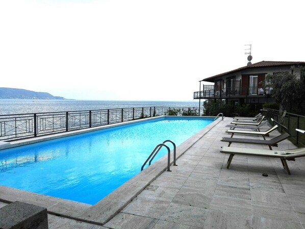 A swimming pool with a view of the sea, a balcony with lounge chairs, and a building with a satellite dish. - Villa Cappellina (Toscolano Maderno)