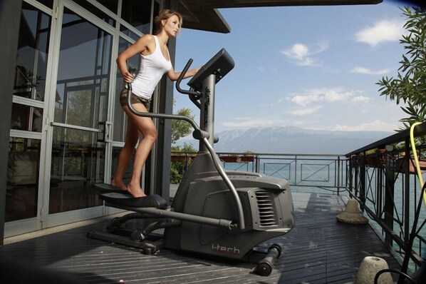 A woman using an elliptical machine on a wooden deck with a glass wall and a view of mountains.