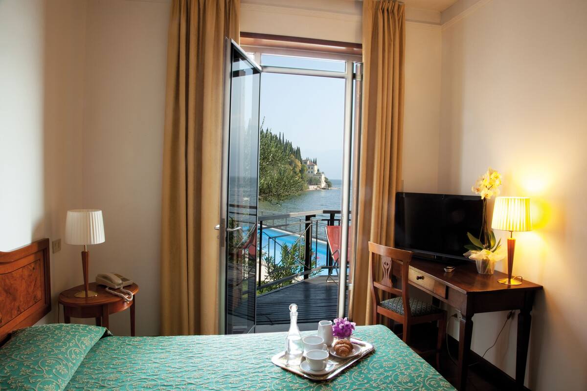 Habitación de hotel con cama, escritorio con televisor, pequeña mesa con bandeja de desayuno y vista a un cuerpo de agua y follaje.