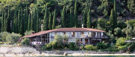 A two-story house with a red roof, located near a body of water with a rocky shoreline.