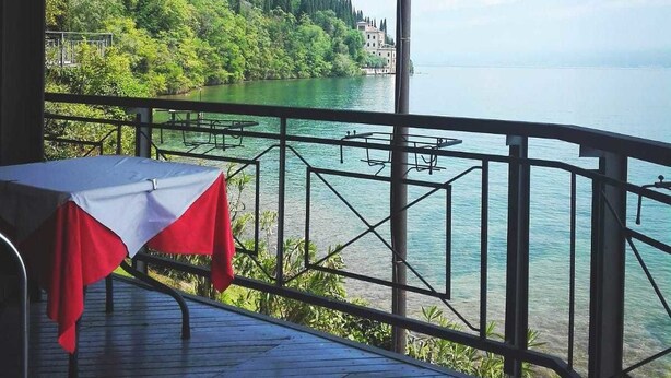A terrace with a table covered by a red and white tablecloth overlooking a lake with buildings and trees in the background. - Villa Cappellina (Toscolano Maderno)
