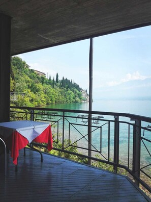 Une terrasse avec une table recouverte d’une nappe rouge et blanche, donnant sur un lac, avec des bâtiments et des arbres en arrière-plan.