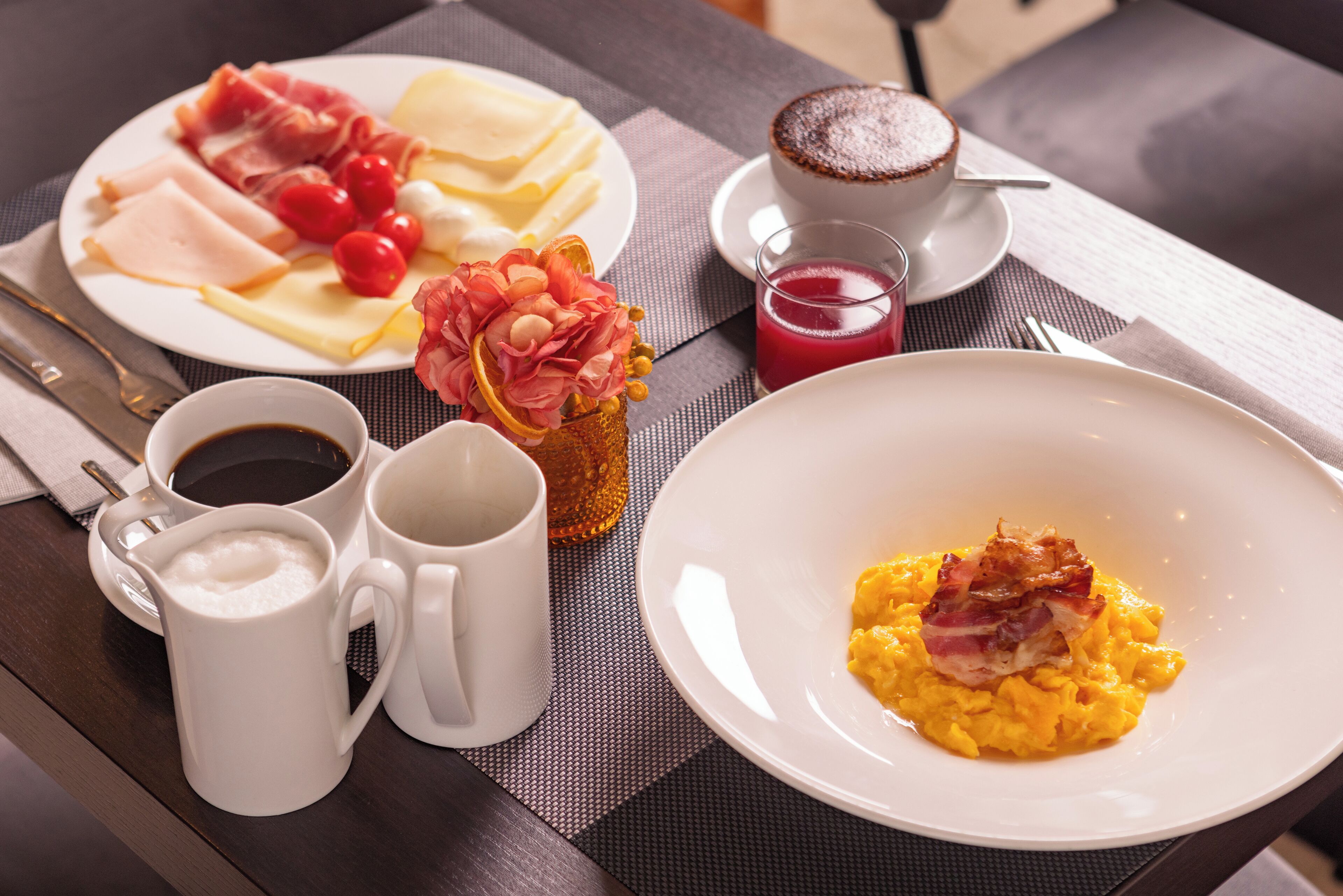 Une table dressée pour le petit-déjeuner, avec une assiette d’œufs brouillés garnis de lardons, une assiette de jambon et de fromage, des tomates cerises et une tasse de café.