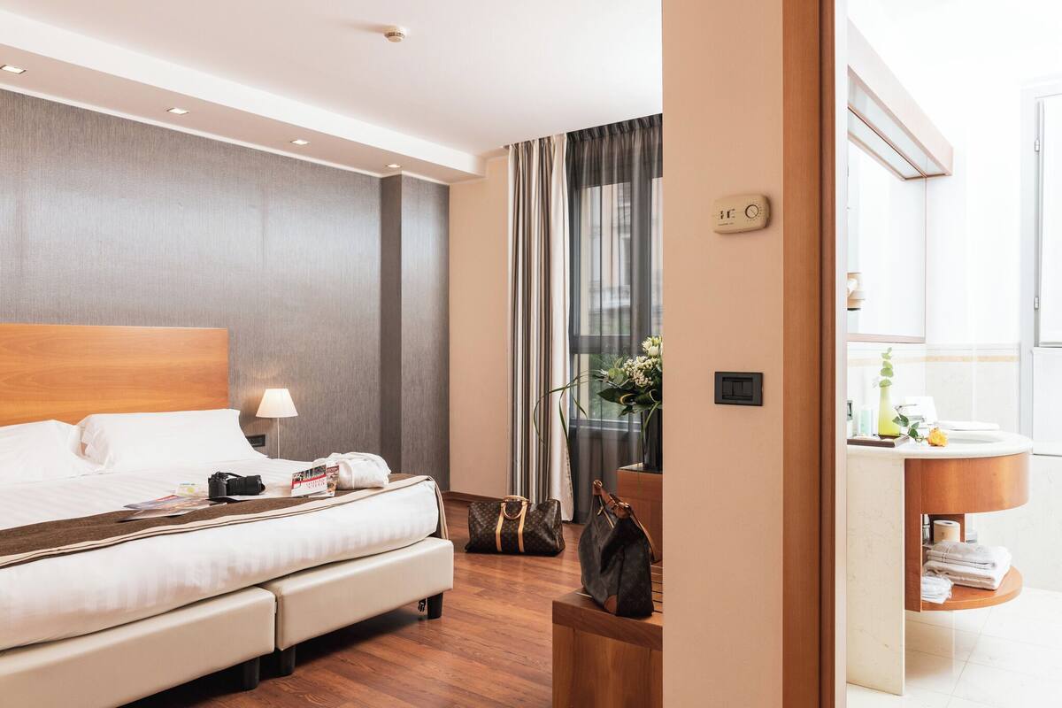 A modern hotel room with a large bed, bedside lamp, and a view of the city through sheer curtains.