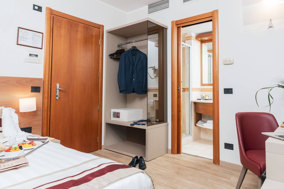 A hotel room with a bed, a desk, a red chair, a wardrobe, and a bathroom visible through an open door.