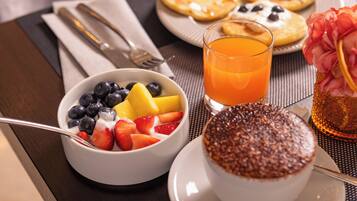 A breakfast setting with a bowl of fruit, a glass of orange juice, pancakes, and a dessert on a table.