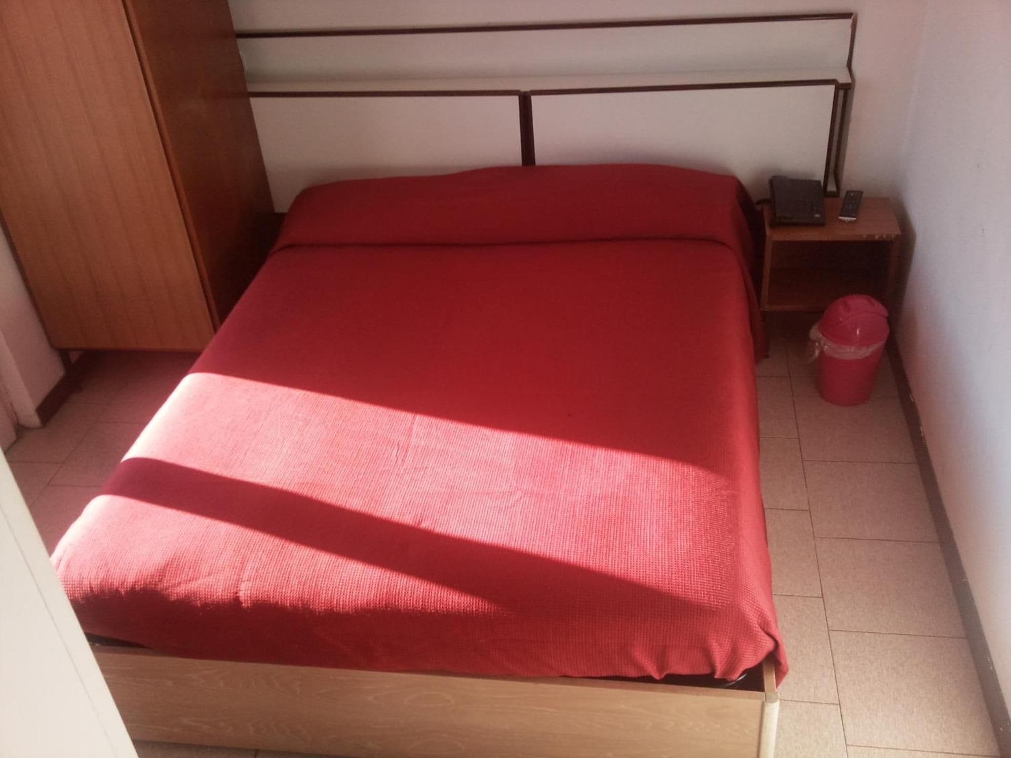 A single bed with a wooden frame and a red mattress in a room with white walls and a tiled floor.