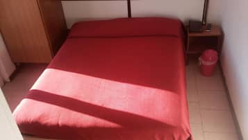 A single bed with a wooden frame and a red mattress in a room with white walls and a tiled floor.
