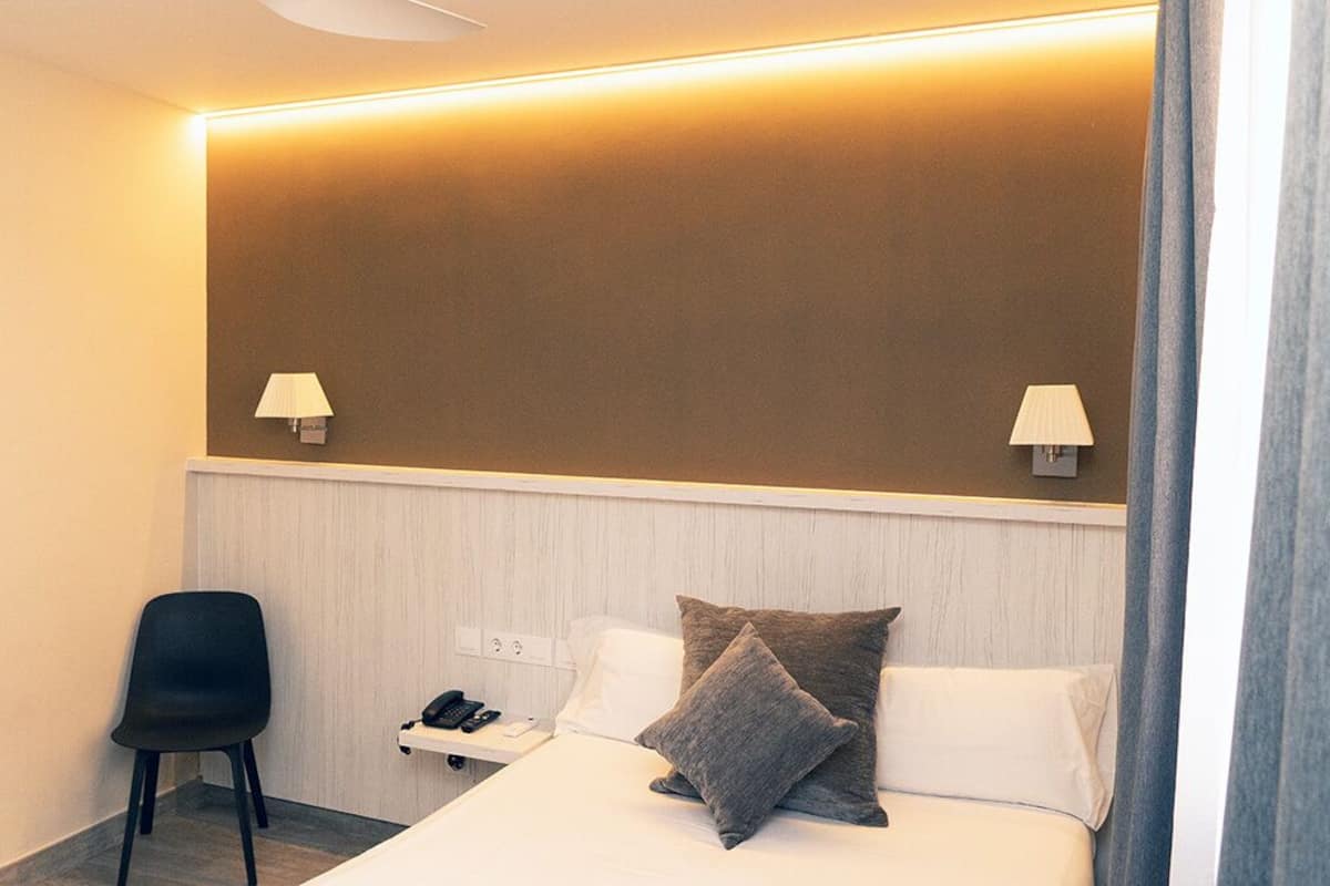 A hotel room with a bed, two bedside lamps, a chair, a bedside table with a phone, and a wall-mounted light.