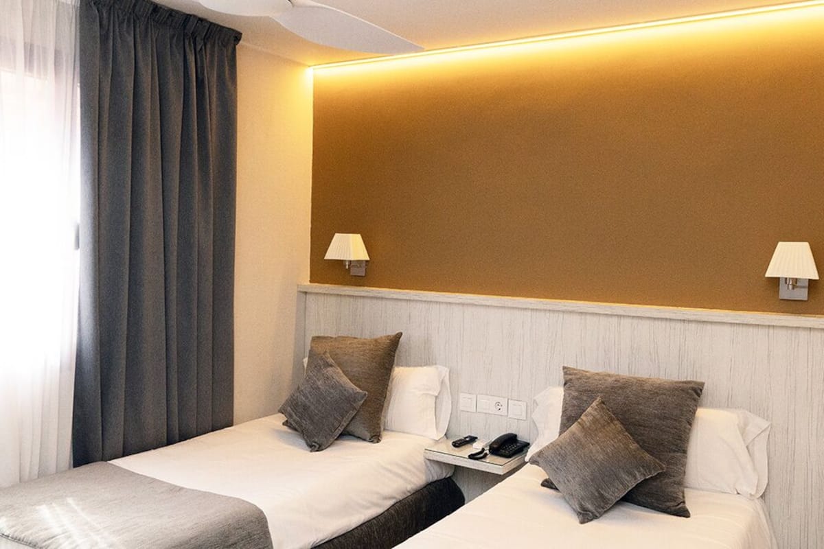 A hotel room with two beds, a bedside table, a lamp, and a wall-mounted phone.