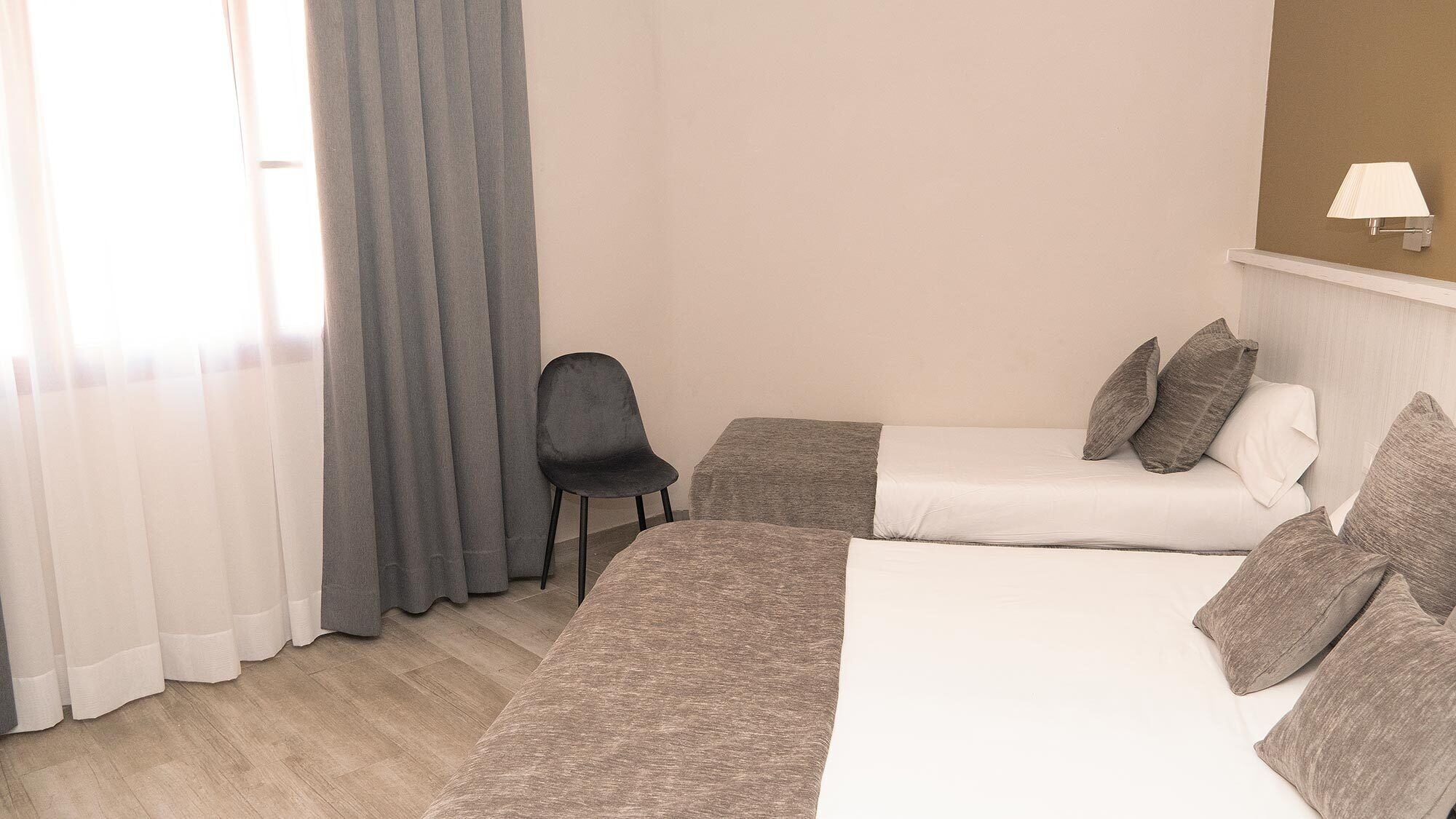 A hotel room with two beds, a chair, a lamp, and a window with curtains.