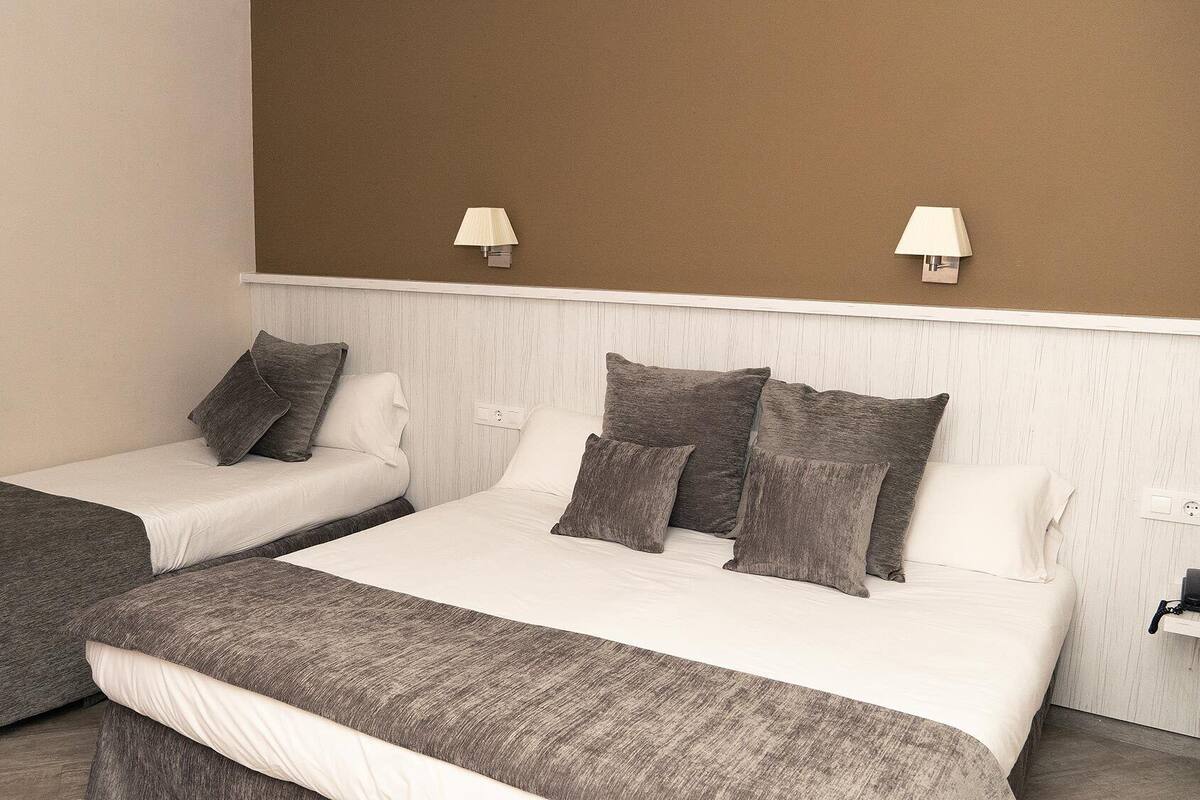 Two single beds with grey and white bedding, a brown wall, and two wall-mounted lamps.