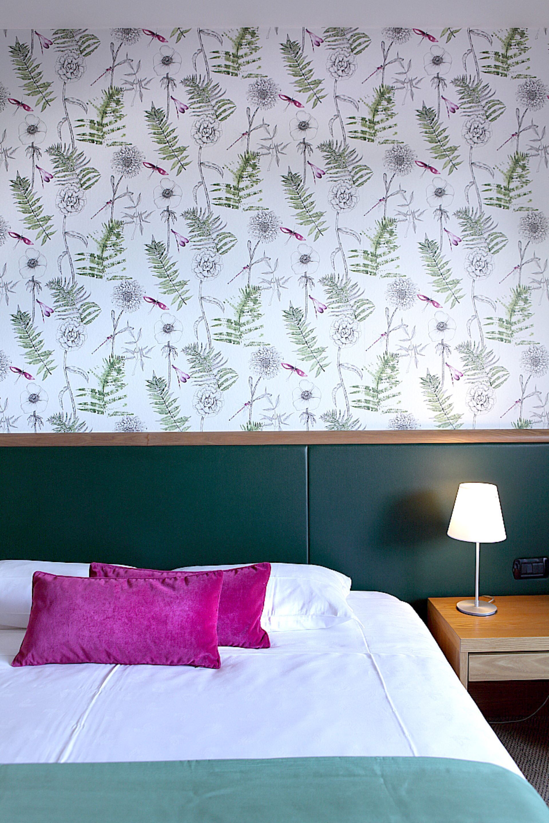 Ein Hotelzimmer mit einem Bett, zwei Kissen, einem Nachttisch mit Lampe und floraler Tapete.