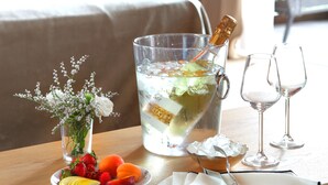 Quarto de hotel com uma cama, uma mesa com um prato de frutas, um jarro de vidro com rolha e dois copos de vinho.