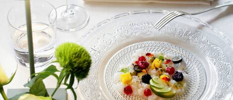 A dining table with a glass plate of food, a wine glass, a small glass of water, a flower, and a napkin with cutlery.