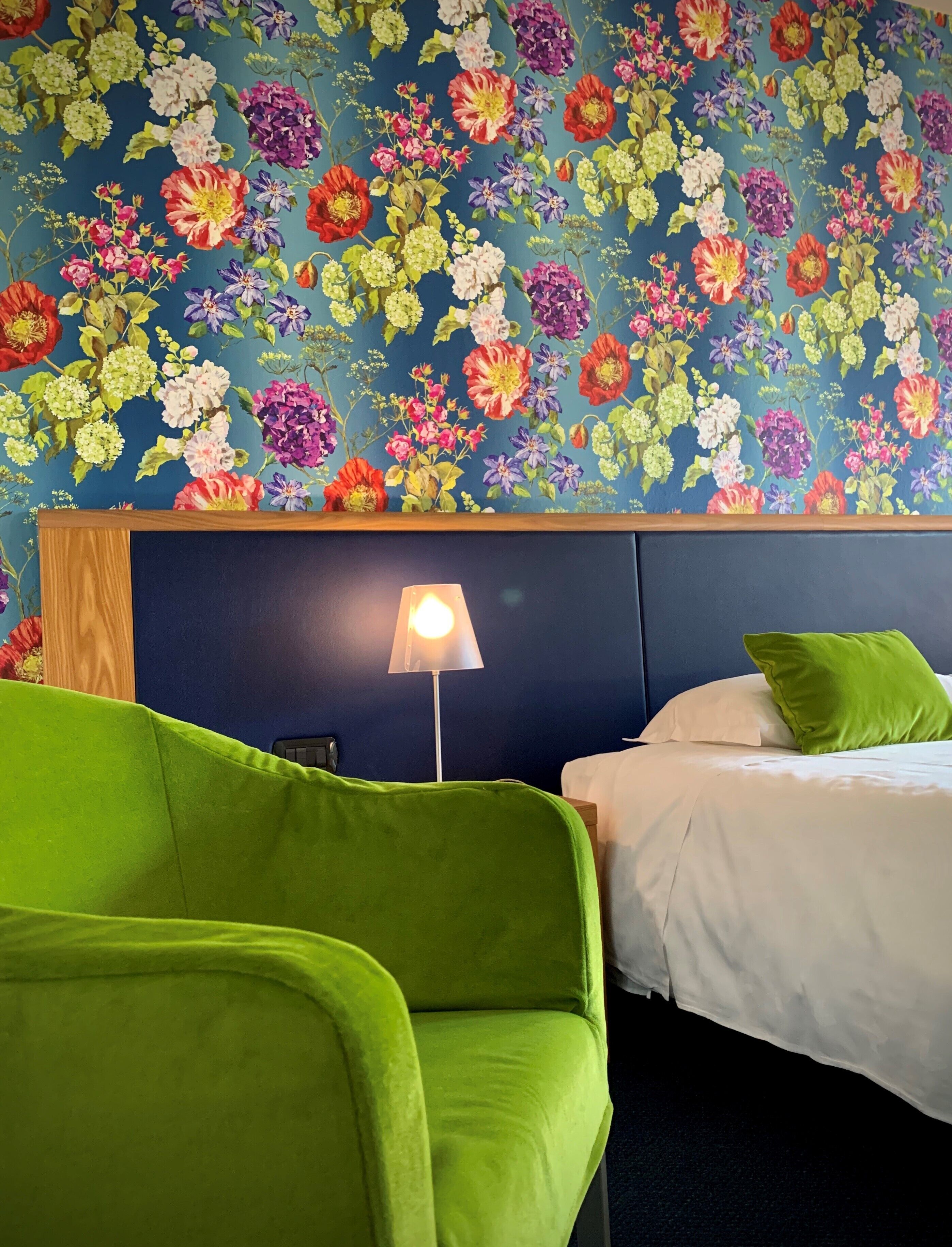 Una camera d'hotel con carta da parati a motivi floreali, una poltrona verde, un comodino con una lampada e un letto con biancheria da letto bianca.