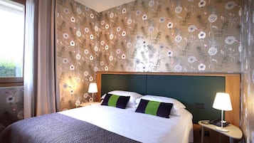 Una habitación de hotel con una cama grande, dos mesitas de noche con lámparas, papel tapiz con motivos florales y una ventana con cortinas.